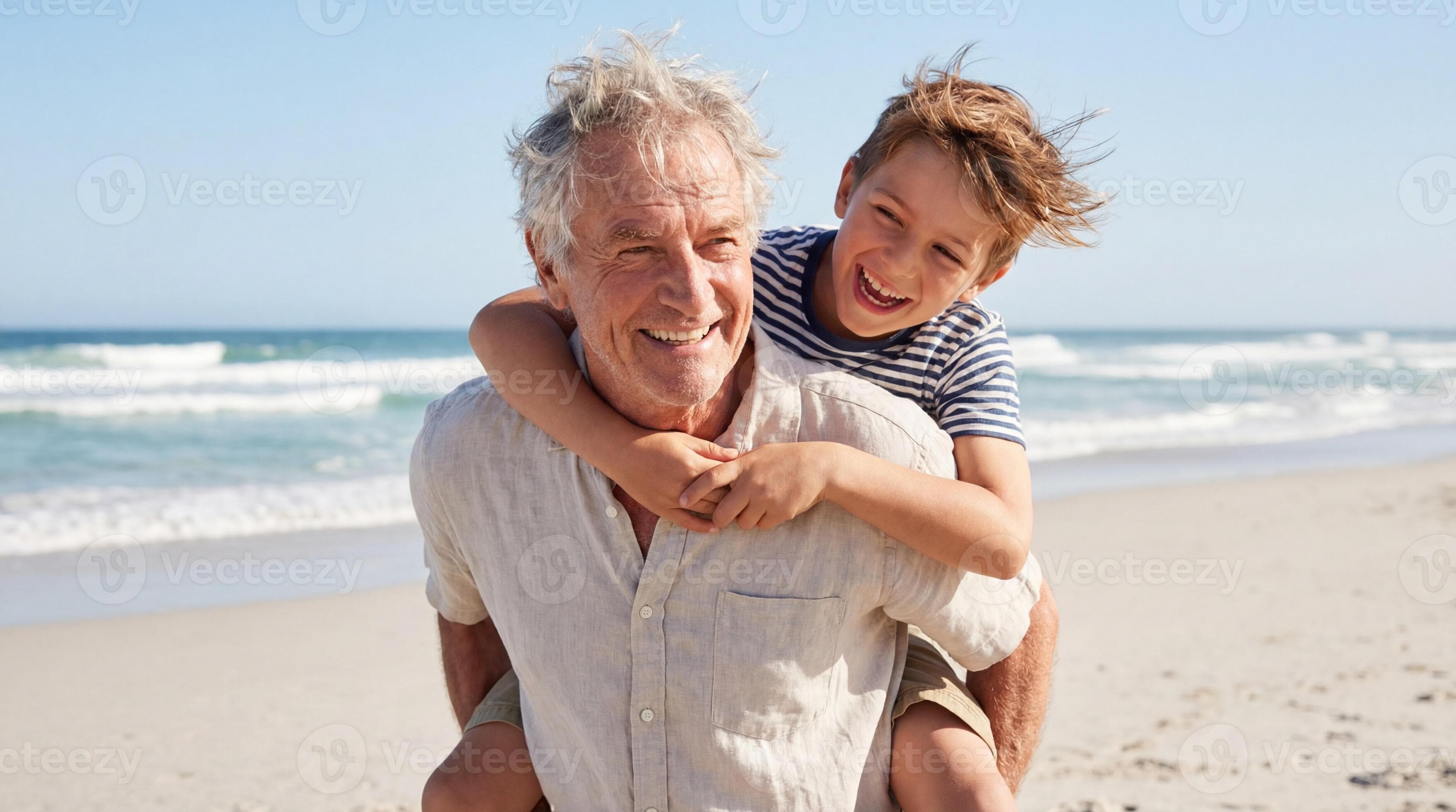 Elderly man carrying young boy on his back while standing on sandy beach with ocean waves and clear blue sky in the background