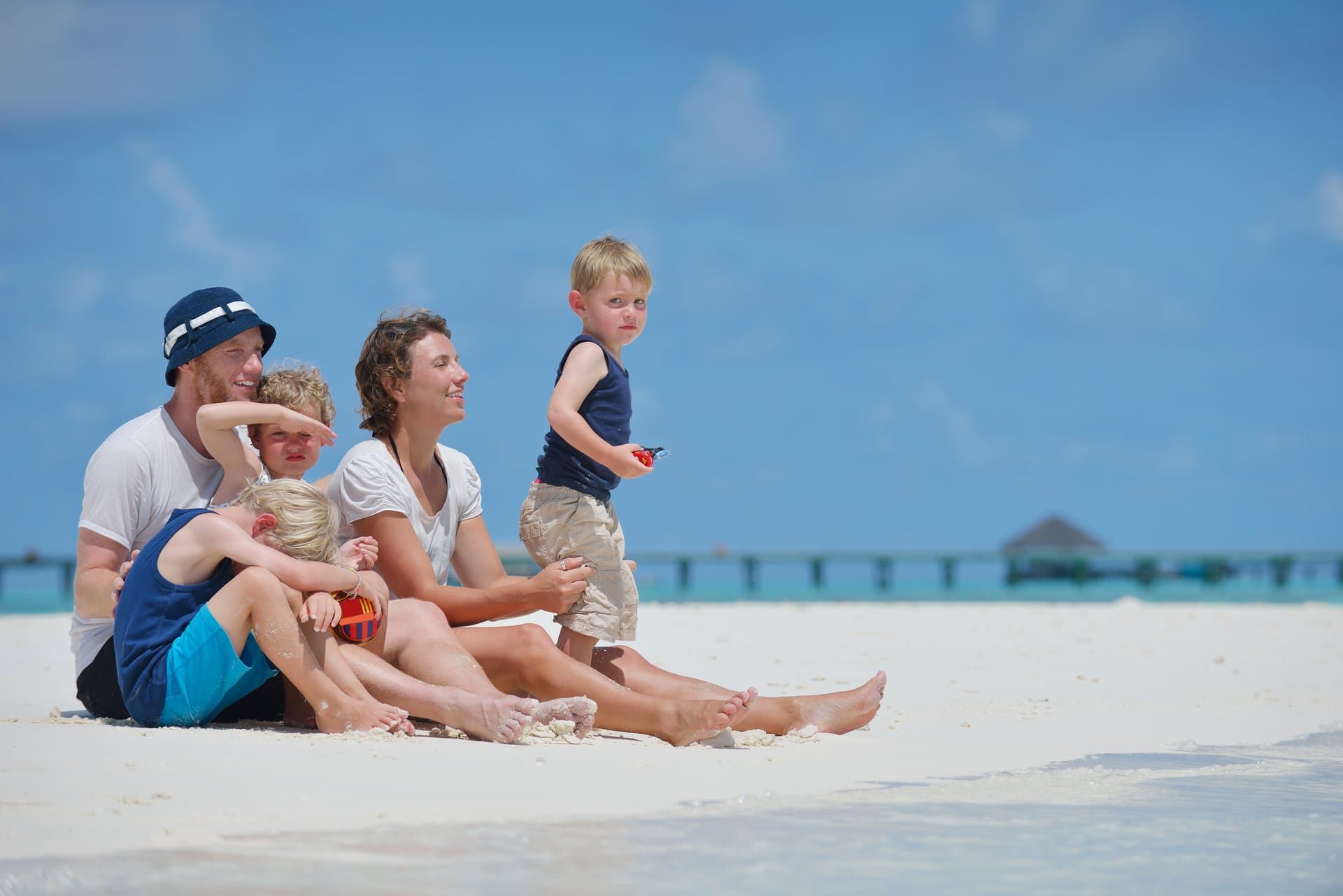 Elderly man carrying young boy on his back while standing on sandy beach with ocean waves and clear blue sky in the background