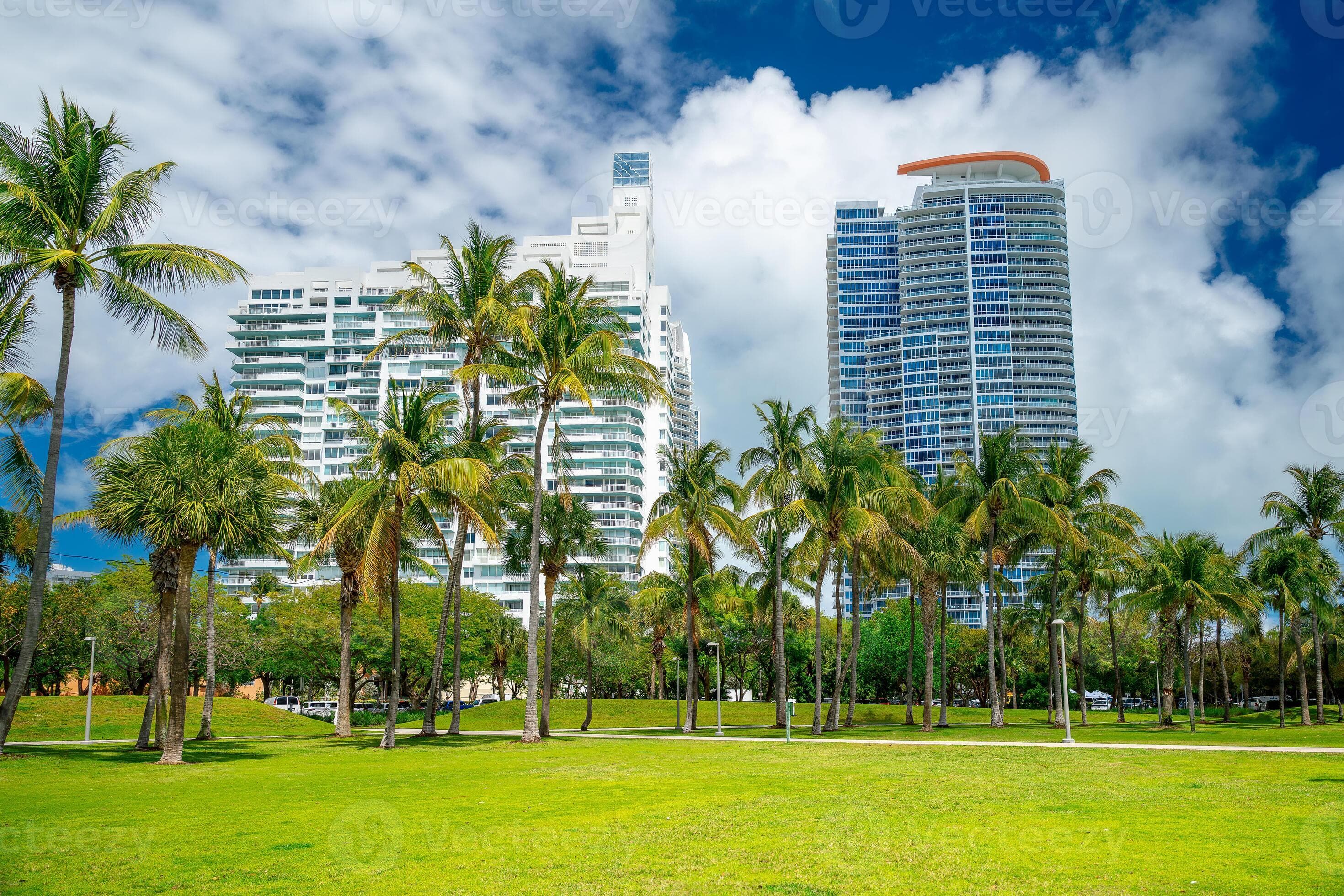 Modern high-rise apartment buildings and tropical park in Miami Beach. Perspective of tall residential towers behind a green field with palm trees under a cloudy sky in Miami.